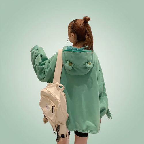 Back view of a woman wearing a cute frog pullover sweatshirt in green with a hood and carrying a beige backpack at night